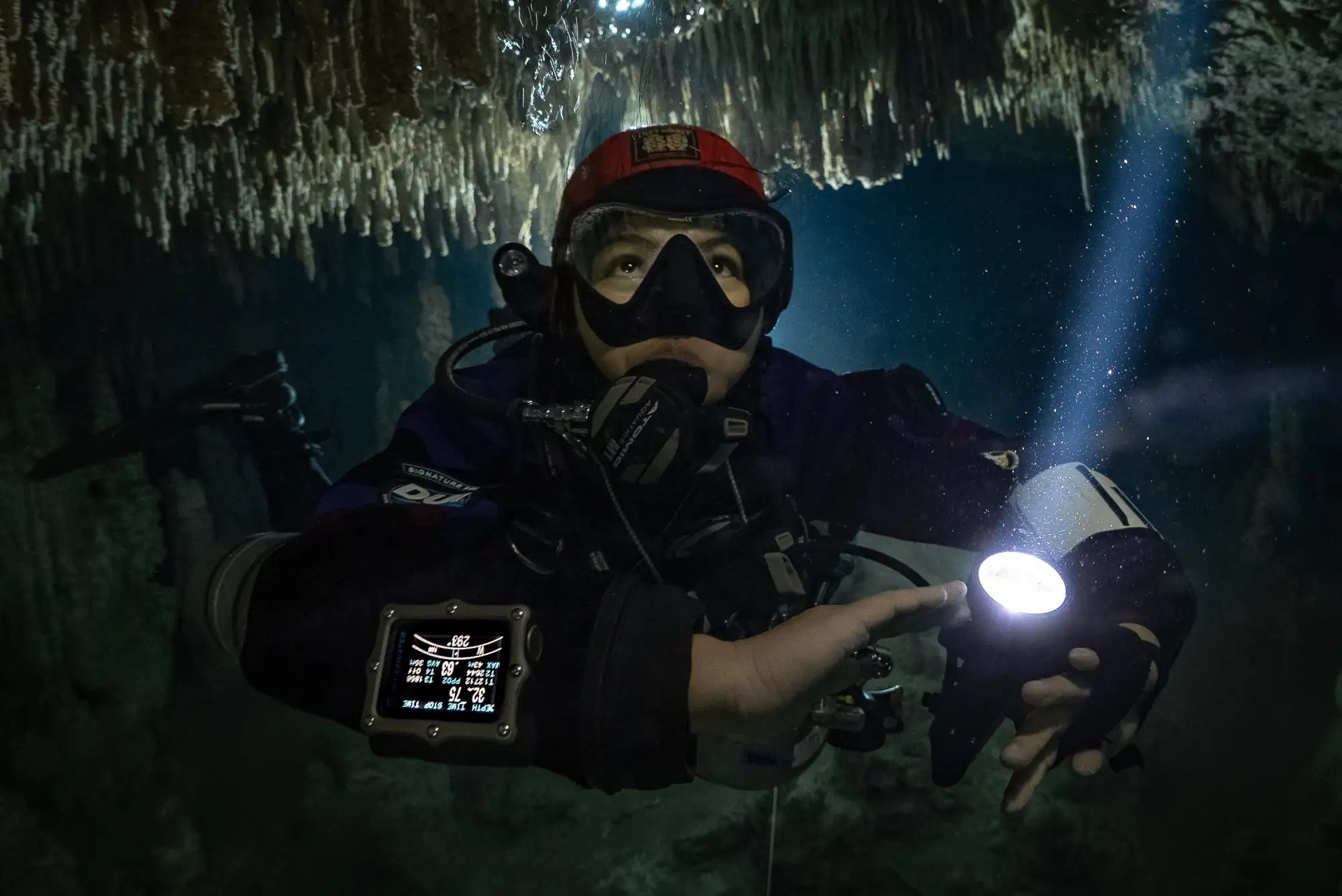 A diver wearing a red helmet and black diving gear holds a bright white flashlight, illuminating the dark underwater cave.
